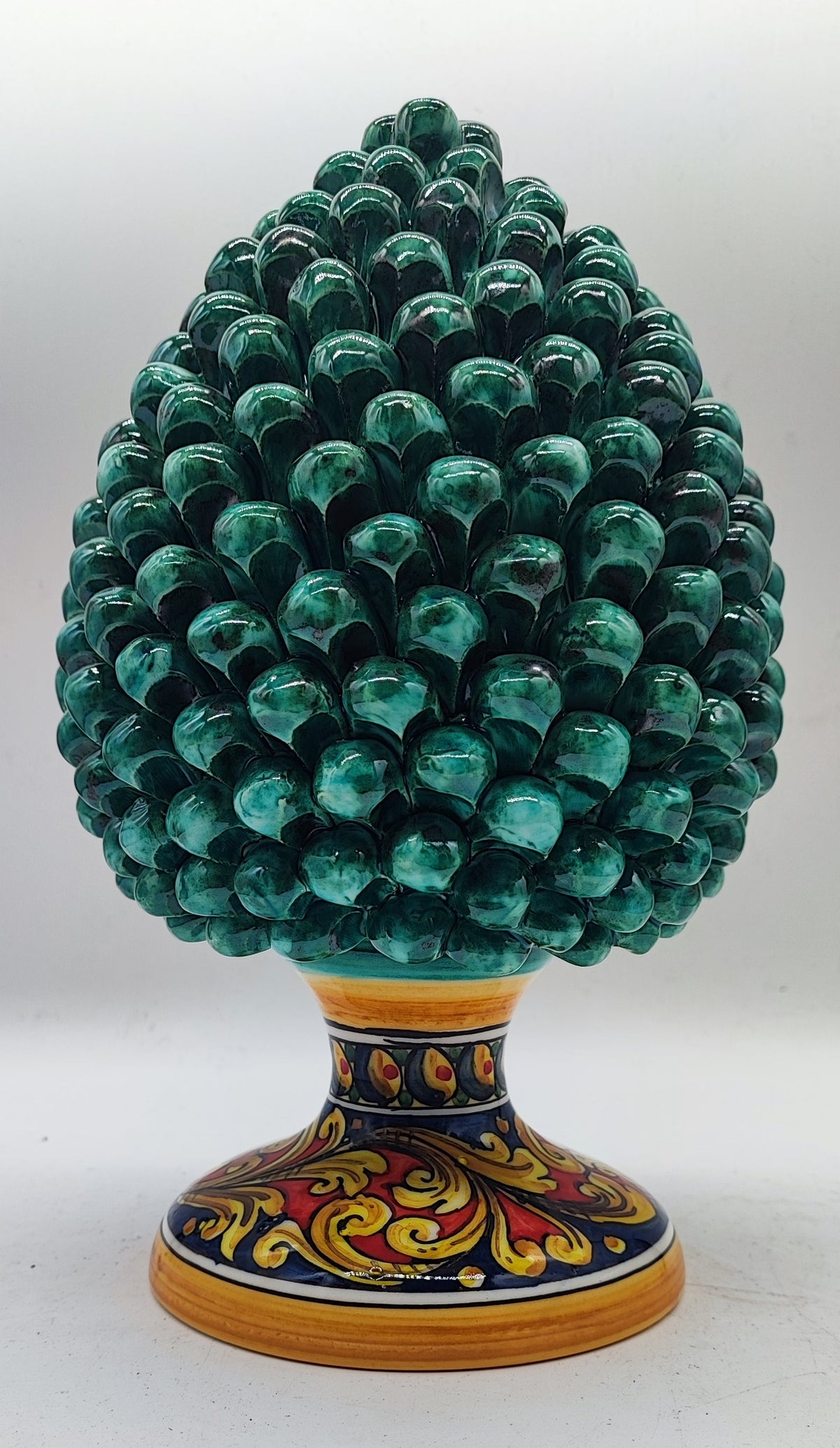 Glossy emerald green Sicilian ceramic pine cone surrounded by Christmas greenery, festive ornaments and warm holiday lights.