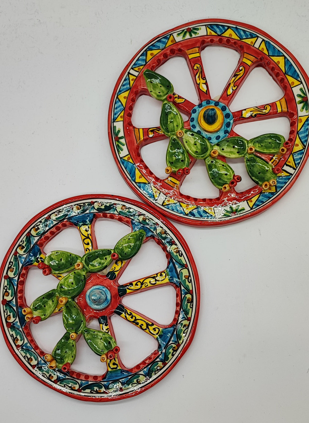 Hands holding a Sicilian ceramic cart wheel with red spokes, blue and yellow patterns and raised prickly pears. Diameter 20 cm, 7.9 inches.