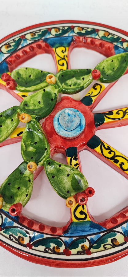 Two colourful ceramic Sicilian cart wheels with raised prickly pears on a wooden table, with hands, paintbrushes and paint pots.