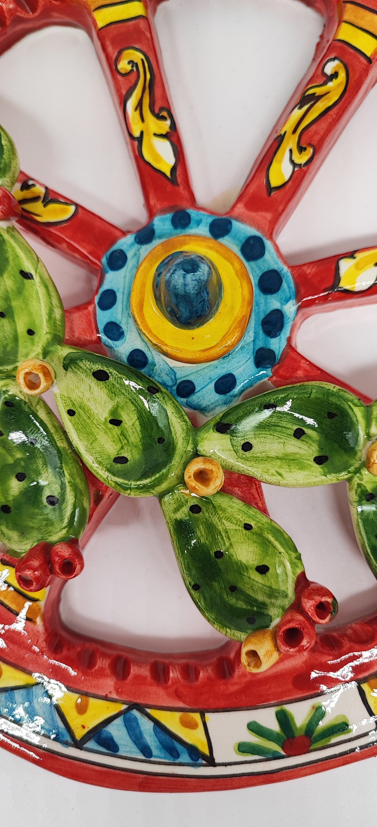 Detail of a ceramic cart wheel with a blue and yellow geometric border, red spokes, yellow scroll patterns and green prickly pears.